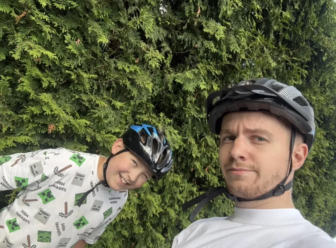 Cyprian w kasku rowerowym robi sobie zdjęcie z uśmiechniętym chłopcem na tle żywopłotu